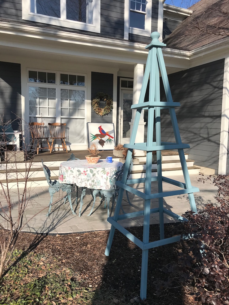 May include: A light blue wooden obelisk stands in a garden setting. The obelisk has a pyramid shape with a square base and is painted in a muted blue color. A small table and chairs are visible in the background.