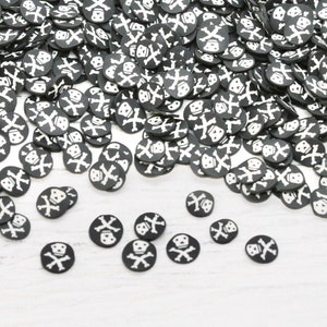 May include: A collection of black and white skull and crossbones polymer clay slices. The circular slices feature a white skull and crossbones design on a black background. These decorative elements are scattered across a white surface.