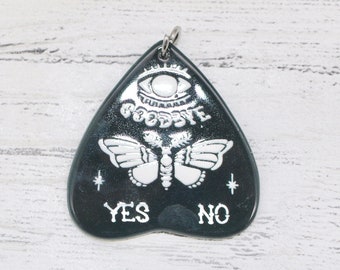 May include: A heart-shaped planchette pendant in black and white. The design includes an eye, a moth, and the words "GOODBYE", "YES", and "NO". A silver-coloured ring is attached at the top.