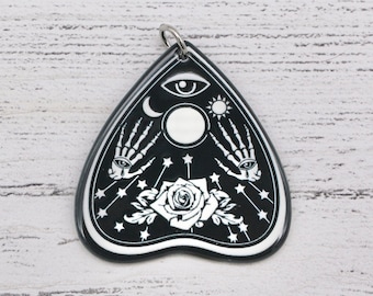 May include: Black and white pendant shaped like a witch's board with a crescent moon, sun, eye, and hands with eyes. The pendant also features a rose and stars.
