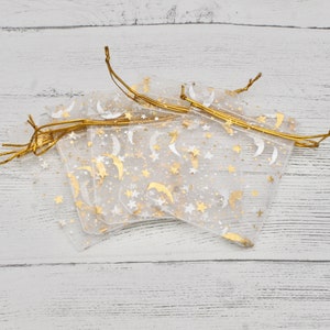 Puede incluir: Un conjunto de cinco bolsas de regalo de organza blanca transparente con lazos de cinta dorada. Las bolsas están decoradas con un patrón de purpurina dorada de estrellas y medias lunas.