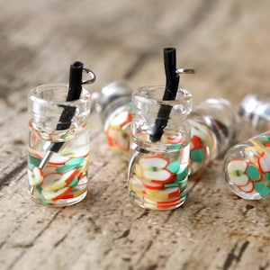 Puede incluir: Cinco frascos de vidrio en miniatura con líquido transparente y rodajas de fruta de colores en su interior. Cada frasco tiene una pajita negra y una tapa plateada con un lazo.