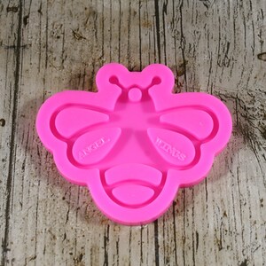May include: Pink silicone mold shaped like a bee with the words "Angel Wings" on the left wing and "Wings" on the right wing.