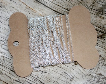 May include: A spool of silver-colored chain, likely for jewelry making, is displayed against a rustic wooden background. The chain is finely linked and appears to be made of metal. The spool is made of brown cardboard.