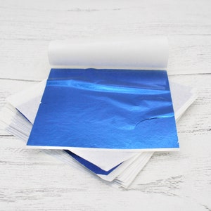 May include: A stack of blue metallic leaf sheets, with one sheet partially unrolled, on a white wooden surface.