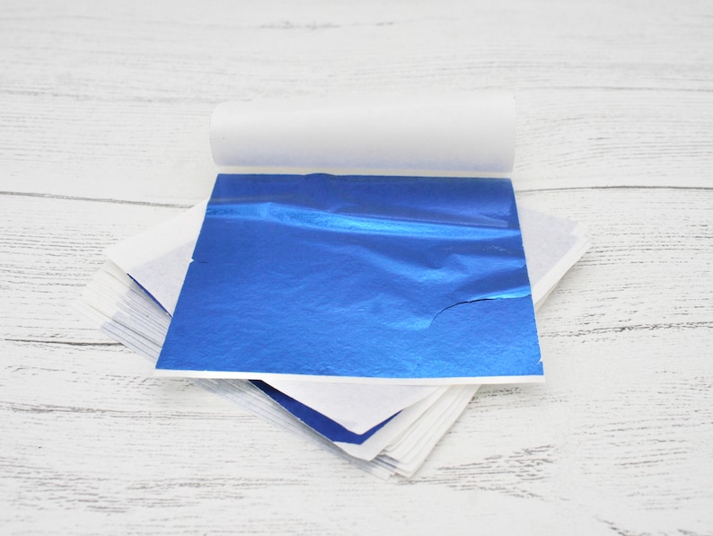 May include: A stack of blue metallic leaf sheets, with one sheet partially unrolled, on a white wooden surface.
