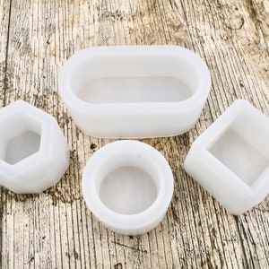 May include: Four white silicone molds for making candles or soap. The molds are in different shapes: oval, round, square, and hexagonal.