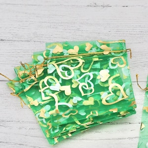 Puede incluir: Un conjunto de bolsas de regalo de organza verde con diseños de corazones dorados. Las bolsas están hechas de tela transparente y tienen un cierre de cordón.