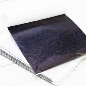 May include: A stack of black metallic leaf sheets, with a single sheet unfolded and lying on top. The sheets have a textured, iridescent finish.