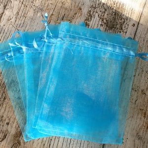 Puede incluir: Un conjunto de cuatro bolsas de regalo de organza azul turquesa con lazos de cinta de satén. Las bolsas están apiladas una encima de la otra y están sobre una superficie de madera.