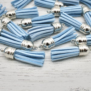 May include: A collection of light blue suede tassels with silver-colored metal caps. The tassels are arranged in a scattered pattern, showcasing their soft texture and the metallic sheen of the caps. Ideal for jewelry making or craft projects.