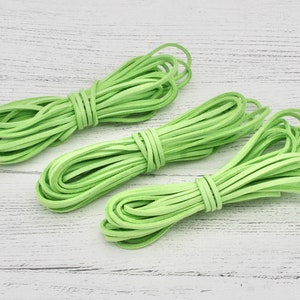 Puede incluir: Tres paquetes de cordón de ante verde claro, cuidadosamente enrollados y atados. Los cordones están dispuestos sobre una superficie de madera blanca envejecida. Los paquetes están espaciados uniformemente, mostrando la textura y el color del material.