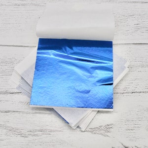 May include: A sheet of blue metallic leafing, folded in half and partially unfolded, revealing the shiny, wrinkled surface. The leafing is on a white background.