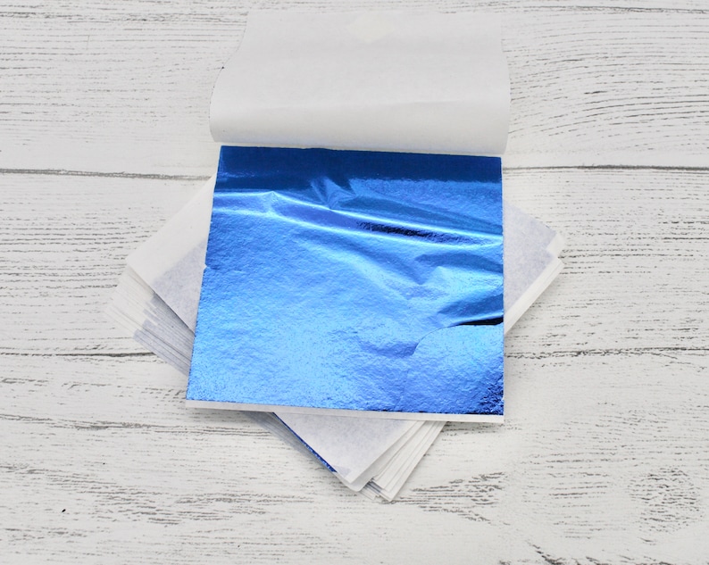 May include: A sheet of blue metallic leafing, folded in half and partially unfolded, revealing the shiny, wrinkled surface. The leafing is on a white background.