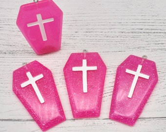 May include: Four pink coffin-shaped charms with silver cross accents. The charms are made of resin and have a sparkly finish.