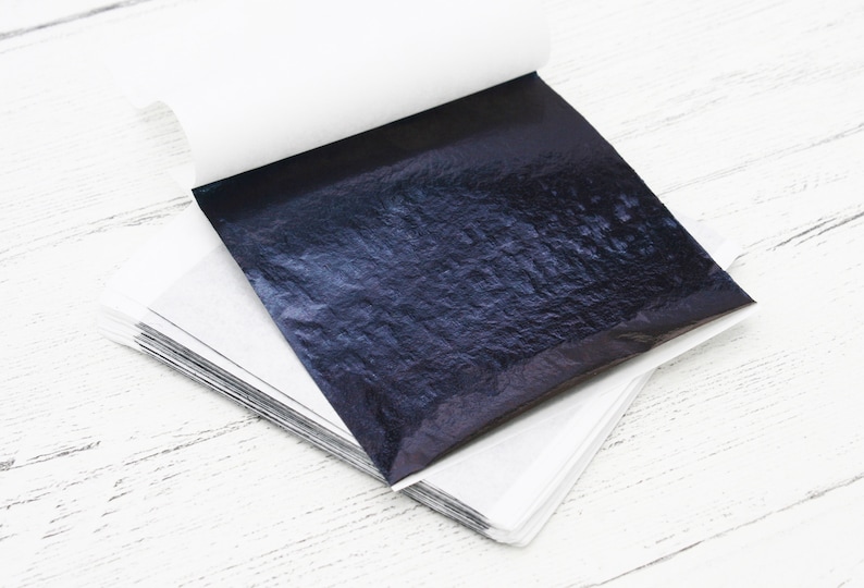 May include: A stack of thin, black, metallic sheets of transfer leaf, with a slightly textured surface, on a white background.