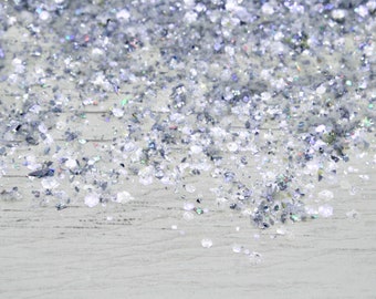 Könnte beinhalten: Nahaufnahme von silbernem Glitter, der auf einer hellgrauen Holzoberfläche verstreut ist. Der Glitter ist schillernd und reflektiert das Licht.
