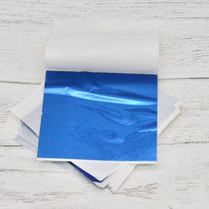 May include: A stack of blue metallic leaf sheets, with a white backing sheet, on a white wooden surface.