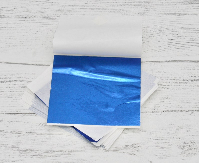 May include: A stack of blue metallic leaf sheets, with a white backing sheet, on a white wooden surface.