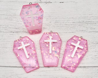 May include: Four pink coffin-shaped resin charms with silver cross accents. The charms have a glittery finish and are perfect for jewelry making or crafting.