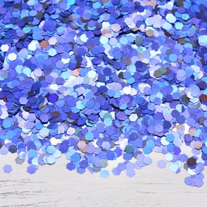 May include: A close-up of a pile of blue glitter. The glitter is made up of many small, hexagonal pieces in various shades of blue, creating a shimmering effect. The glitter is scattered on a white surface.