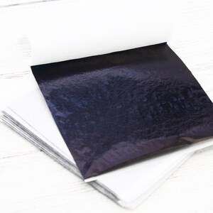 May include: A stack of black metallic leaf sheets, each sheet individually wrapped in white paper. The sheets are on a white wooden surface.