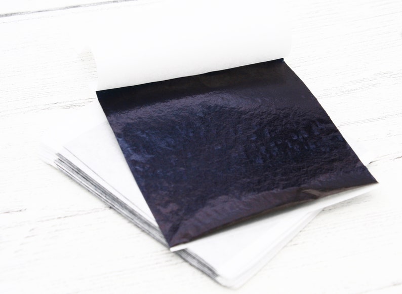 May include: A stack of black metallic leaf sheets, each sheet individually wrapped in white paper. The sheets are on a white wooden surface.