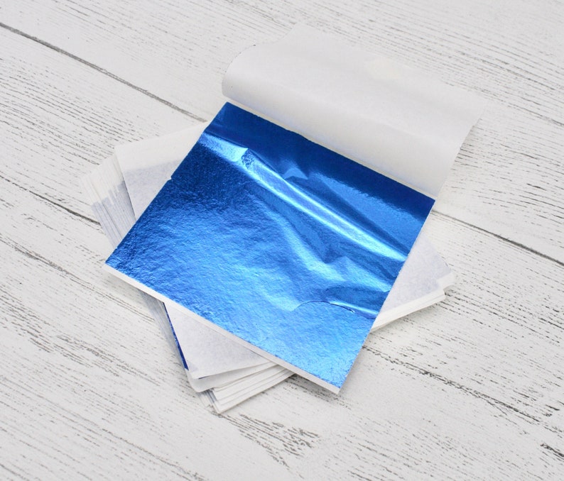 May include: A stack of blue gold leaf sheets, with one sheet partially unfolded, revealing the shiny, metallic surface. The sheets are packaged in a white paper envelope.