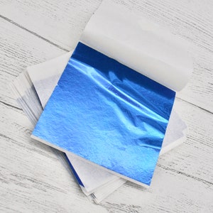 May include: A stack of blue metallic leaf sheets, each sheet is encased in white tissue paper. The sheets are on a white wooden surface.