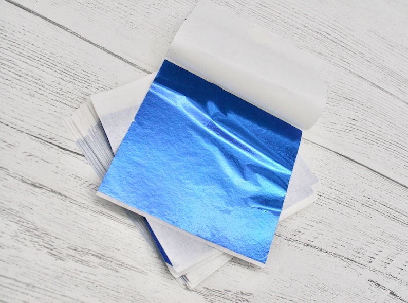 May include: A stack of blue metallic leaf sheets, each sheet is encased in white tissue paper. The sheets are on a white wooden surface.