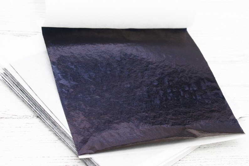 May include: A stack of thin, black, metallic sheets of transfer leaf. The sheets are on a white surface.