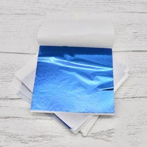 May include: A stack of blue metallic leaf sheets, with a white paper backing, on a white wooden surface.