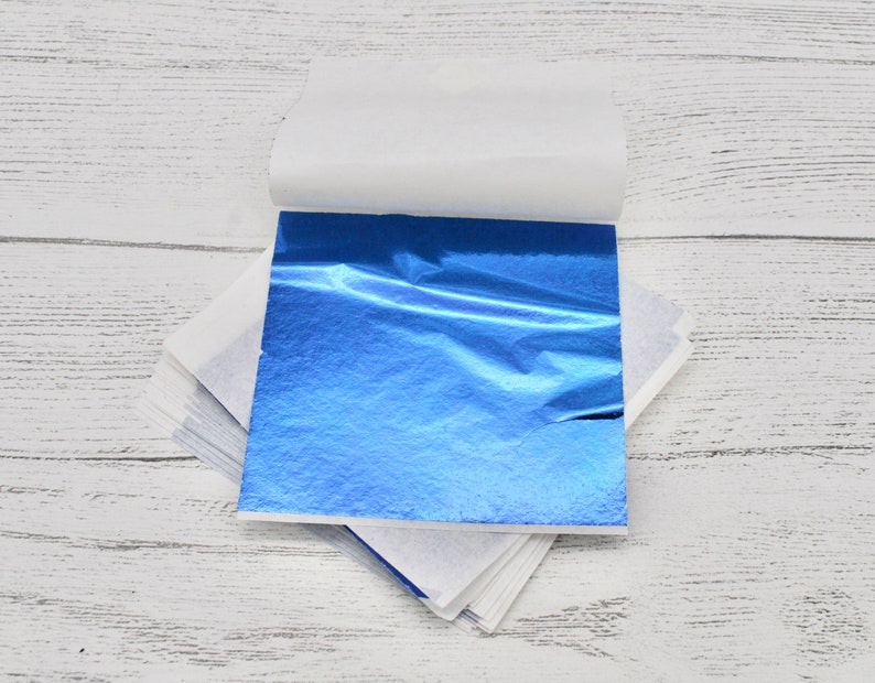 May include: A stack of blue metallic leaf sheets, with a white paper backing, on a white wooden surface.