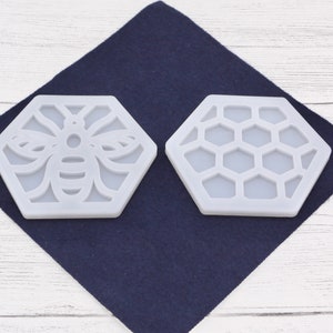 Può includere: Due stampi in silicone bianco a forma di esagono. Uno stampo presenta un design a forma di ape, mentre l'altro ha un motivo a nido d'ape.