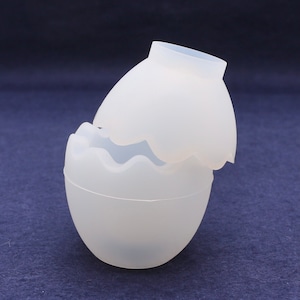 Può includere: Uno stampo in silicone bianco a forma di uovo con un bordo ondulato. Lo stampo è aperto e la metà superiore è appoggiata sulla metà inferiore.