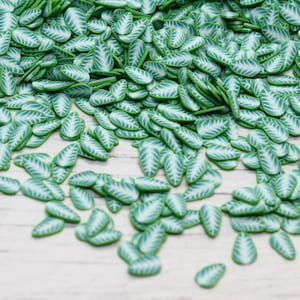 May include: A large quantity of small, green and white leaf-shaped polymer clay charms. The charms are scattered on a white wooden surface.