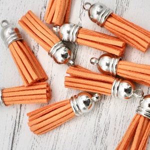 May include: Close-up of several orange suede tassels with silver-colored metal caps. The tassels are arranged on a light-colored wooden surface. Each tassel has a loop for attaching to a project. These are ideal for jewelry making and craft projects.