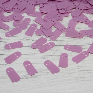 May include: A scattering of purple ghost-shaped confetti on a white wooden surface. The confetti is made of paper or cardstock and is perfect for Halloween decorations.