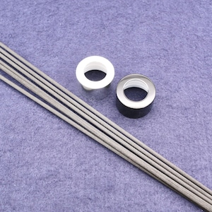 Puede incluir: Dos tapones de botella redondos, uno negro y uno plateado, con un agujero en el centro. Un juego de palitos difusores grises están sobre una superficie morada.