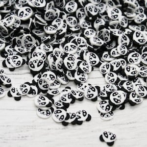 May include: A large pile of black and white panda face shaped resin embellishments. The embellishments are scattered on a white wooden surface.