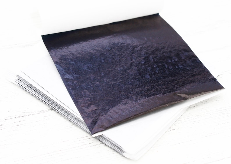 May include: A stack of thin, black, metallic sheets of transfer leaf. The sheets are square and have a slightly textured surface.