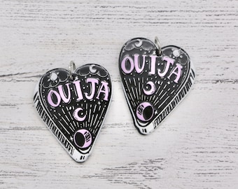 May include: Two heart-shaped Ouija board pendants. The pendants are black with white and light purple accents. The word "OUJIA" is written in light purple. The design includes moons, stars, and a planchette. Each pendant has a silver loop at the top.