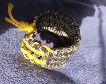 Può includere: Un braccialetto intrecciato con un motivo mimetico e accenti gialli. Il braccialetto è realizzato in paracord e ha un nodo all'estremità.