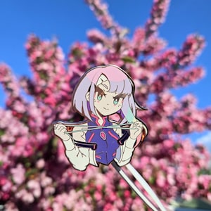 Könnte beinhalten: Eine Metall-Emaille-Anstecknadel mit einer Anime-Figur mit kurzem rosa und hellviolettem Haar, die eine dunkelblaue und weiße Jacke trägt und ein Paar Essstäbchen horizontal vor der Brust vor einem klaren blauen Himmel hält.