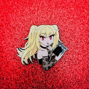 May include: A pin featuring an anime-style character with blonde hair, red eyes, and a finger to the lips. The character is holding a black book with Japanese text. The pin has a black choker and black sleeves with a rose pattern.