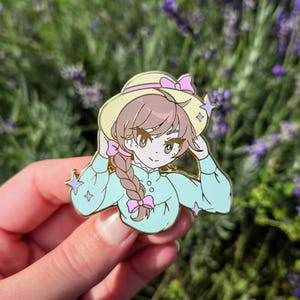 Puede incluir: Un pin de esmalte azul claro con una niña de dibujos animados con cabello castaño en una trenza, usando un sombrero de paja amarillo con una cinta rosa, una camisa azul claro con un lazo rosa y una chaqueta azul claro. El pin tiene un acabado brillante y está colocado sobre un fondo borroso de flores de lavanda.