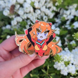 May include: A gold-coloured enamel pin featuring an anime character with orange hair, wearing a red and black suit with the number "02" on the chest. The character is smiling and has large blue eyes.