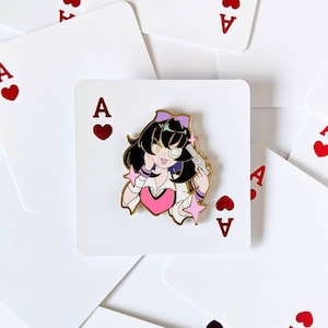 May include: A gold-colored enamel pin featuring a cartoon character with black hair, a pink shirt, and a gun pointed at their head. The character is wearing a white headband with a purple bow. The pin is set against a white background with red hearts and the letter 'A' printed on it.