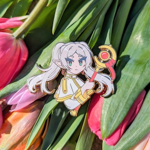 May include: A white enamel pin featuring a cartoon character with long white hair, wearing a white and gold outfit and holding a red and gold staff with a crescent moon and a red gem. The character has green eyes and pink blush.