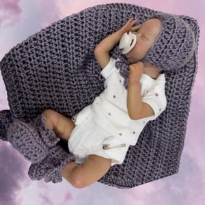May include: A baby doll wearing a white onesie with colorful polka dots, a purple crocheted hat, and matching booties. The doll is lying on a purple crocheted blanket.